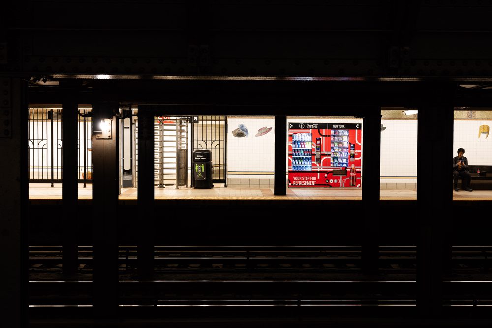 View across the tracks at the 23rd Street-Broadway subway station: an automated retail machine lit within the tiled platform wall, framed by steel columns